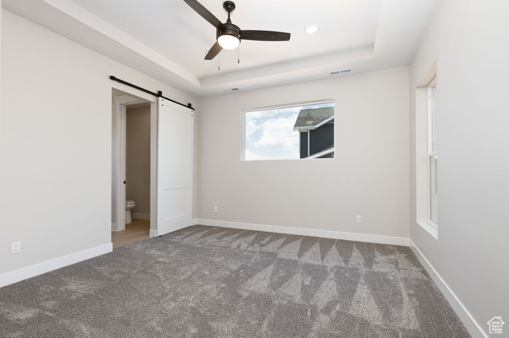 Unfurnished bedroom with a tray ceiling, a barn door, carpet flooring, and a ceiling fan