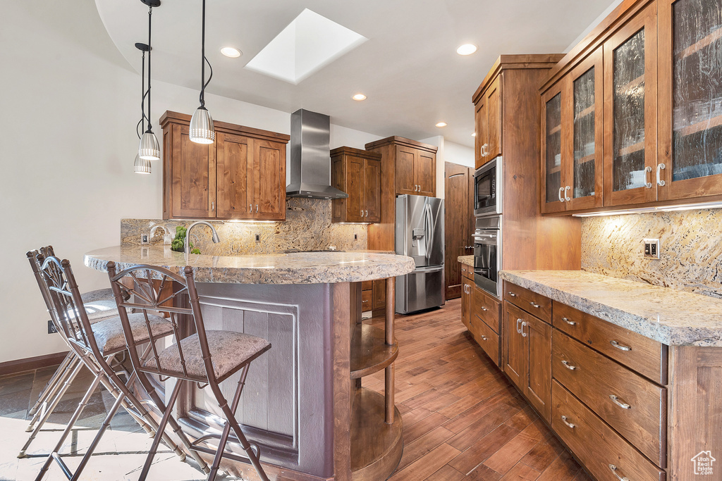 Kitchen featuring a kitchen breakfast bar, a peninsula, a skylight, appliances with stainless steel finishes, and wall chimney exhaust hood