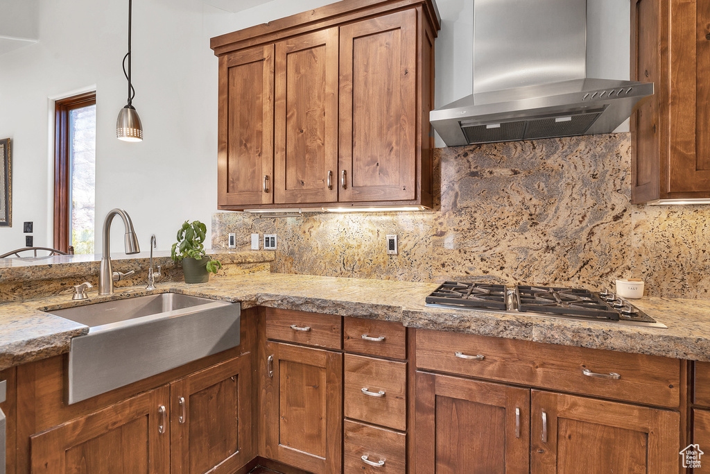 Kitchen with decorative backsplash, brown cabinets, stainless steel gas stovetop, wall chimney exhaust hood, and a sink