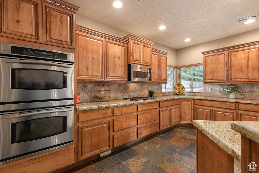Kitchen featuring stone tile floors, decorative backsplash, brown cabinets, and stainless steel appliances
