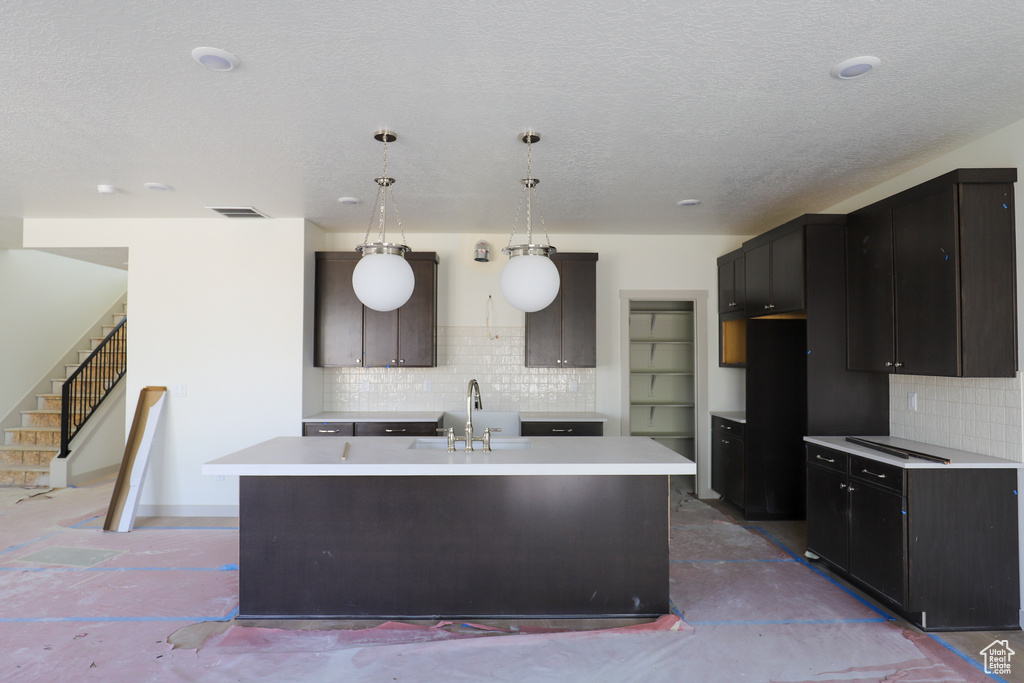 Kitchen with a textured ceiling, visible vents, a kitchen island with sink, a sink, and backsplash