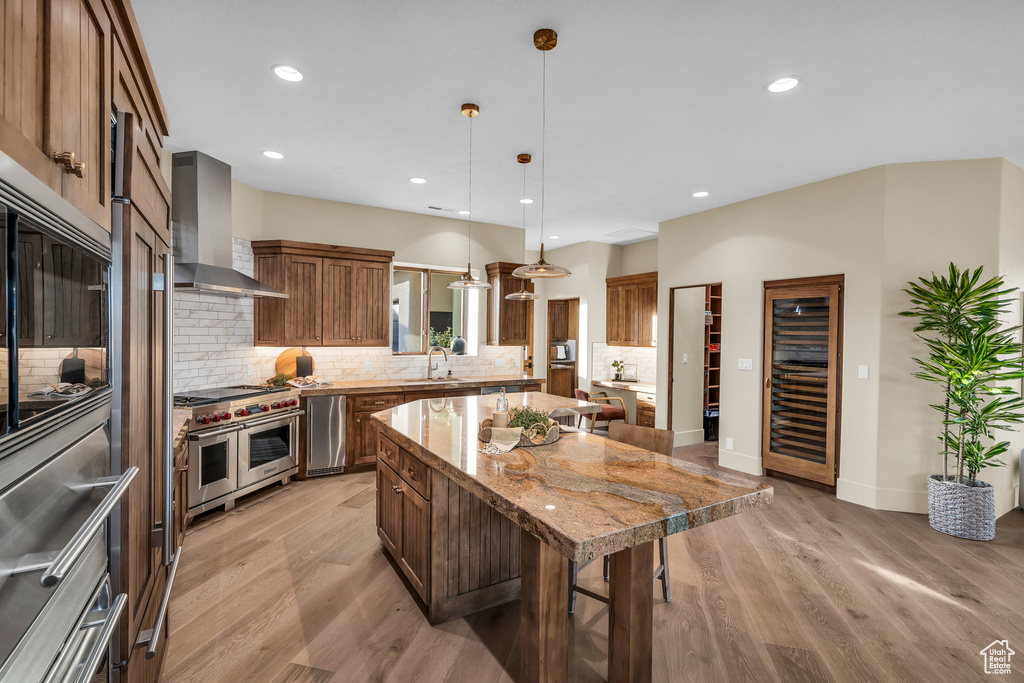Kitchen with a breakfast bar, light wood-style flooring, a kitchen island, appliances with stainless steel finishes, and wall chimney exhaust hood