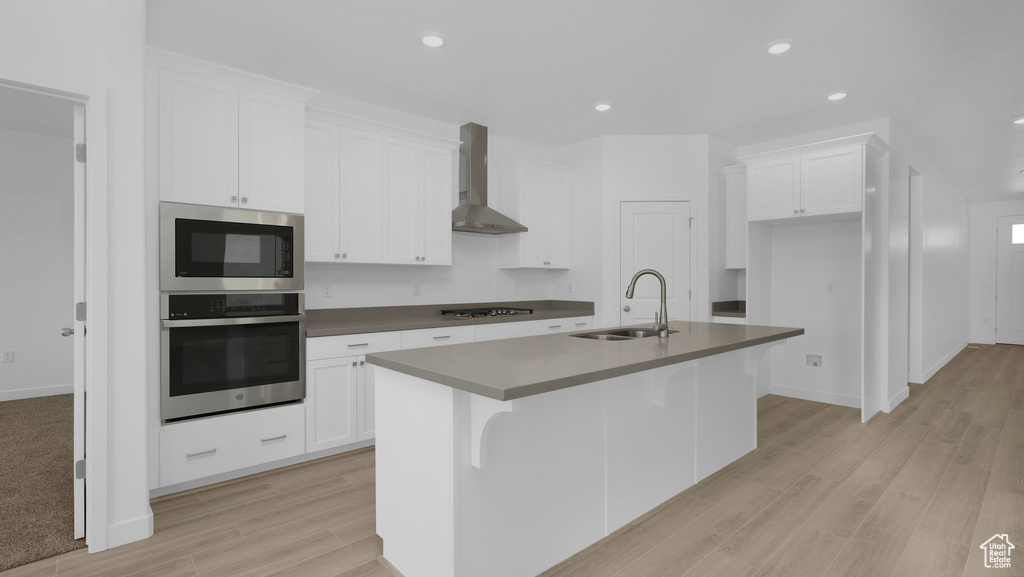 Kitchen featuring appliances with stainless steel finishes, wall chimney exhaust hood, a sink, a kitchen island with sink, and white cabinetry