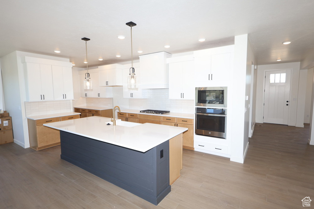 Kitchen with light wood finished floors, an island with sink, a sink, decorative backsplash, and appliances with stainless steel finishes