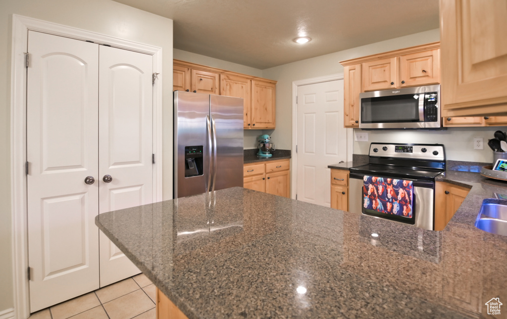 Kitchen featuring light brown cabinets, dark stone countertops, stainless steel appliances, a peninsula, and light tile patterned floors