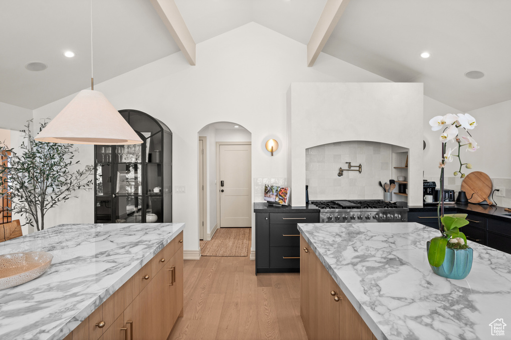 Kitchen with light wood-type flooring, arched walkways, light stone countertops, stove, and vaulted ceiling with beams