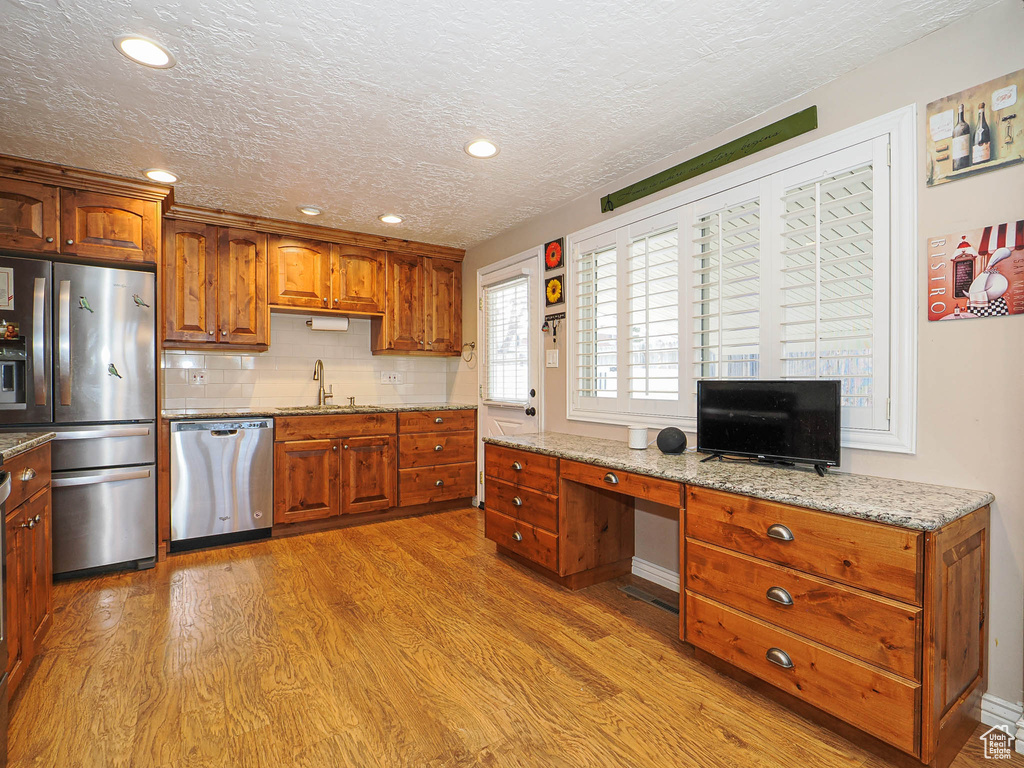 Kitchen with light stone counters, brown cabinetry, decorative backsplash, appliances with stainless steel finishes, and light wood-type flooring