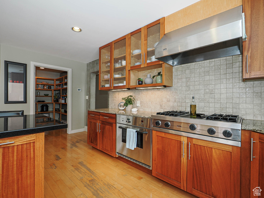 Kitchen with brown cabinetry, stainless steel appliances, light wood-style floors, and exhaust hood