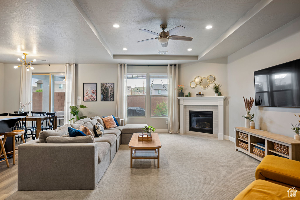 Carpeted living room featuring a tiled fireplace, ceiling fan with notable chandelier, a tray ceiling, recessed lighting, and baseboards