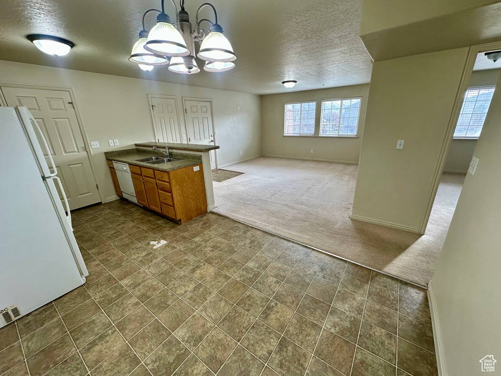 Kitchen featuring white appliances, a peninsula, an inviting chandelier, brown cabinetry, and dark colored carpet