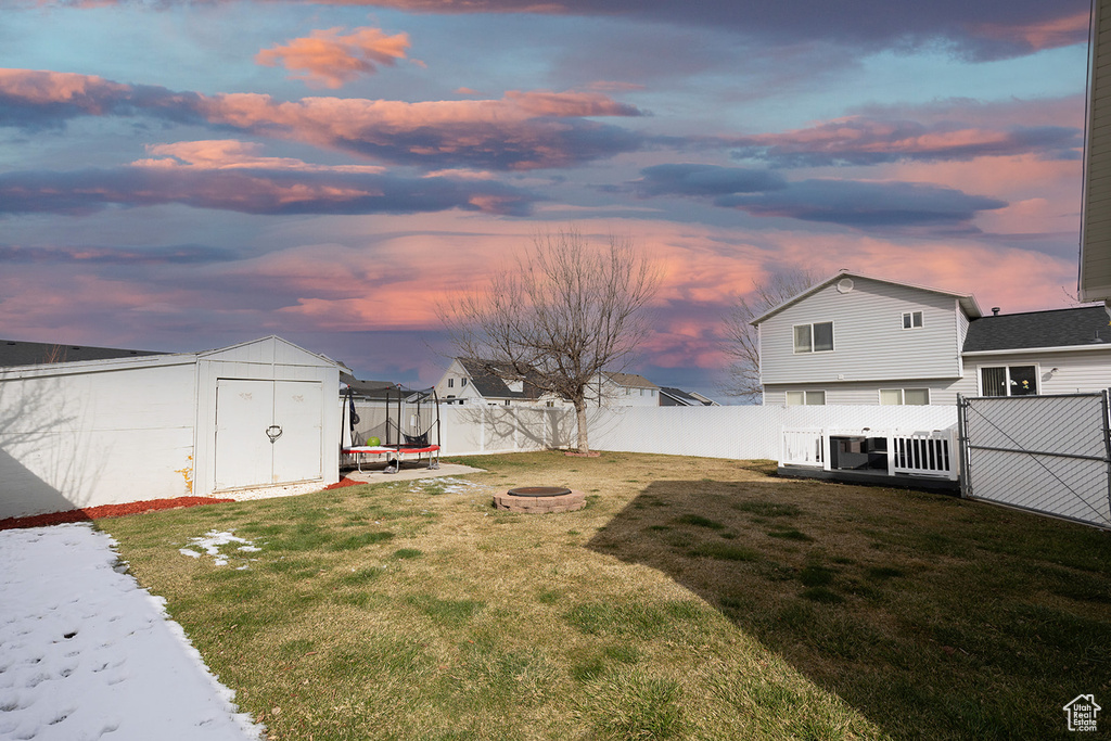 Yard at dusk with an outbuilding, a fenced backyard, a storage unit, a fire pit, and a trampoline