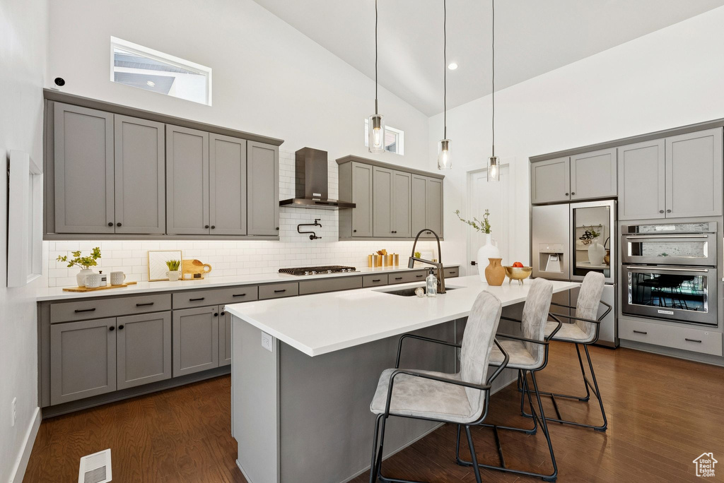 Kitchen featuring gray cabinetry, high vaulted ceiling, wall chimney range hood, appliances with stainless steel finishes, and tasteful backsplash