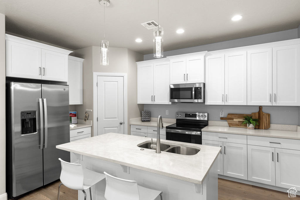 Kitchen with appliances with stainless steel finishes, a sink, white cabinets, hanging light fixtures, and an island with sink