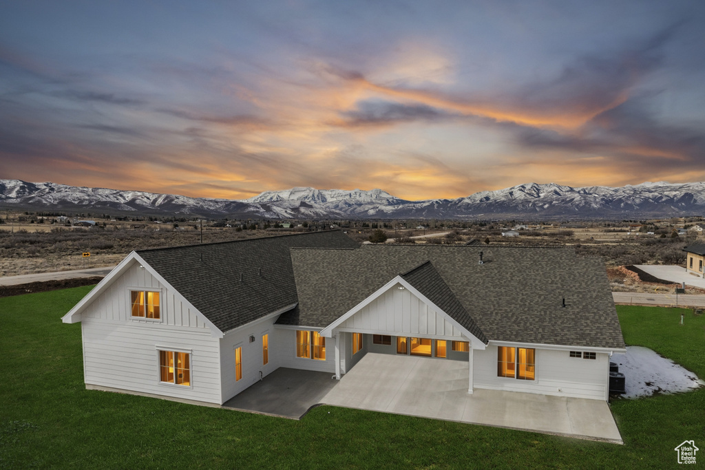 Back of property at dusk featuring a patio, board and batten siding, roof with shingles, a lawn, and a mountain view