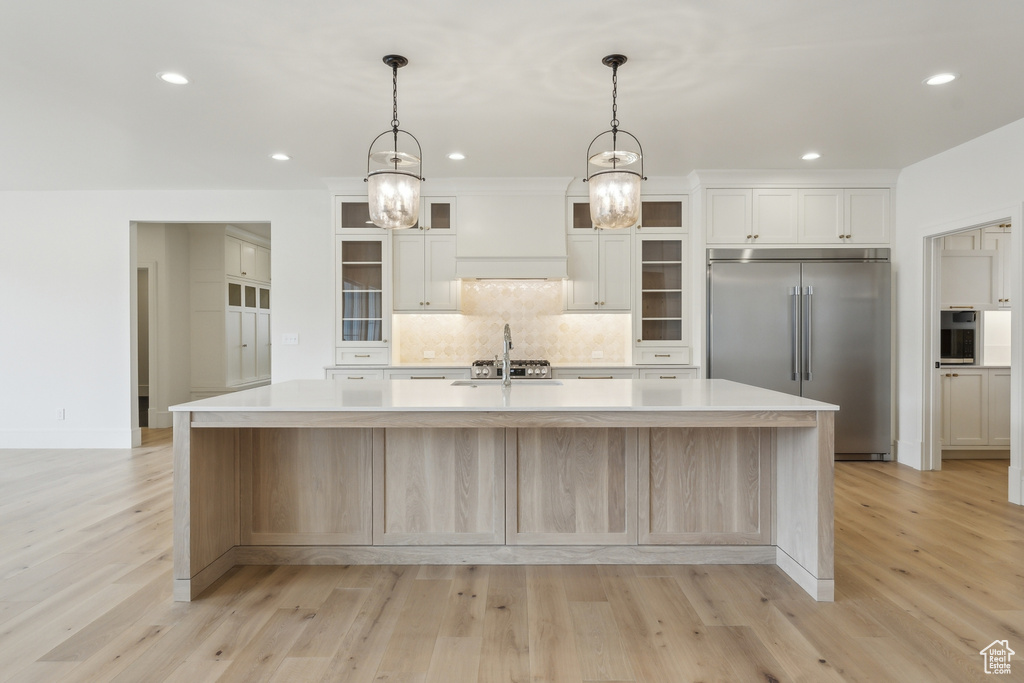 Kitchen featuring stainless steel built in fridge, a large island with sink, light wood-type flooring, custom range hood, and decorative backsplash