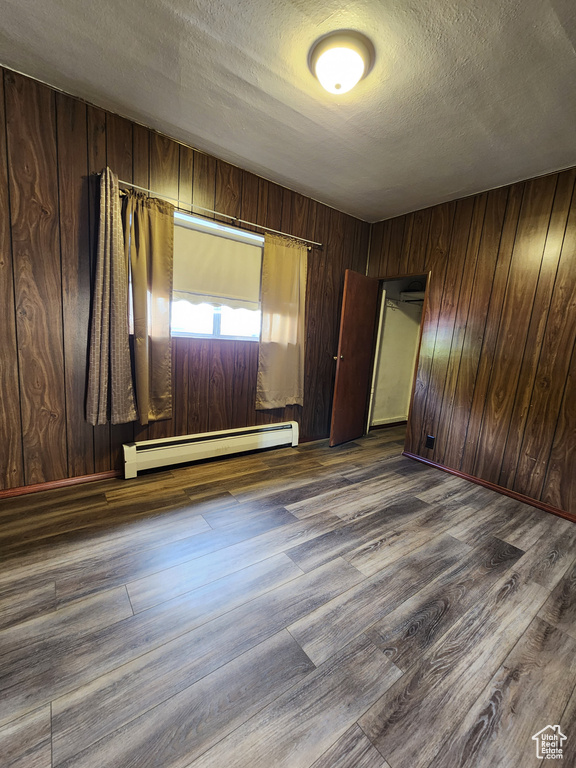 Empty room featuring a baseboard radiator, dark wood-style flooring, a textured ceiling, and wooden walls