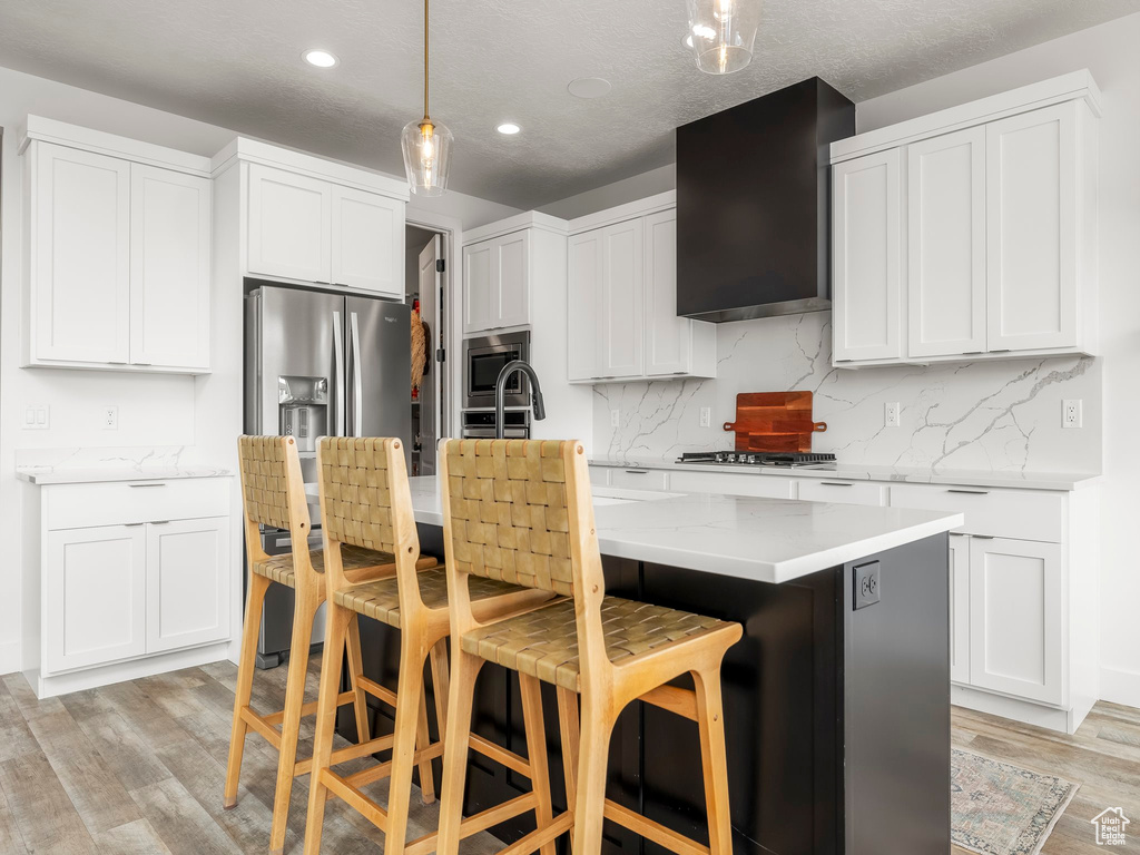 Kitchen with a center island with sink, light wood finished floors, wall chimney exhaust hood, and appliances with stainless steel finishes