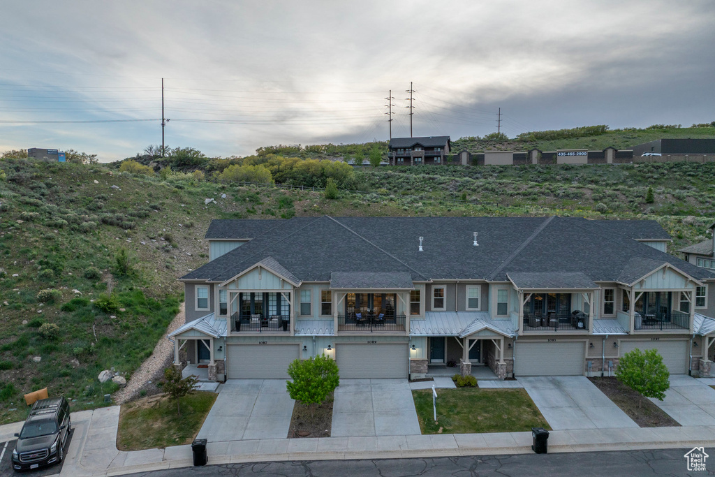 View of property featuring a balcony, driveway, an attached garage, stone siding, and a shingled roof