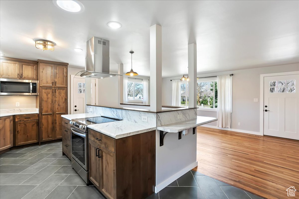 Kitchen featuring a kitchen breakfast bar, island range hood, light tile patterned floors, stainless steel appliances, and brown cabinetry