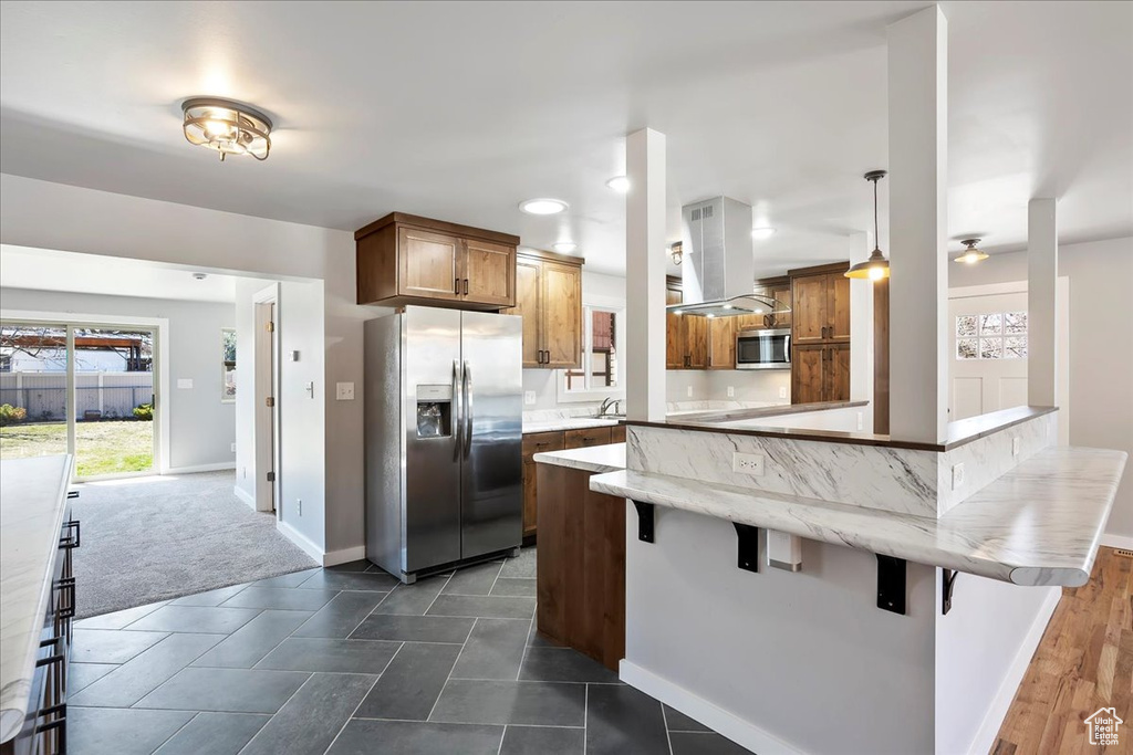 Kitchen featuring a kitchen bar, appliances with stainless steel finishes, a peninsula, brown cabinets, and island range hood