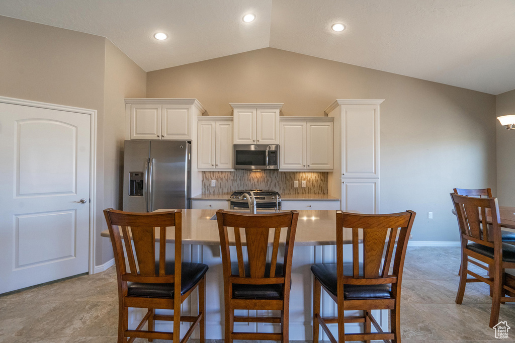 Kitchen with vaulted ceiling, backsplash, appliances with stainless steel finishes, a center island with sink, and white cabinets