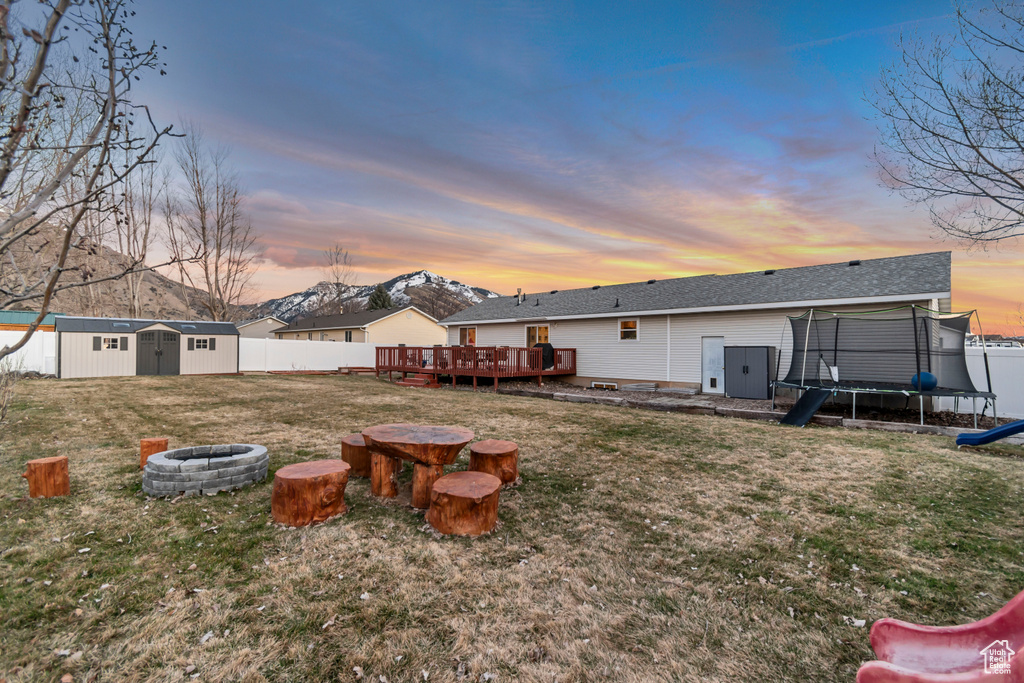Rear view of house featuring a deck with mountain view, a storage shed, a trampoline, a fenced backyard, and a fire pit