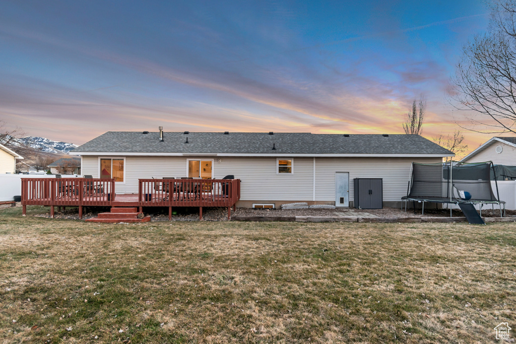 Back of house at dusk with a trampoline, a wooden deck, roof with shingles, and a lawn