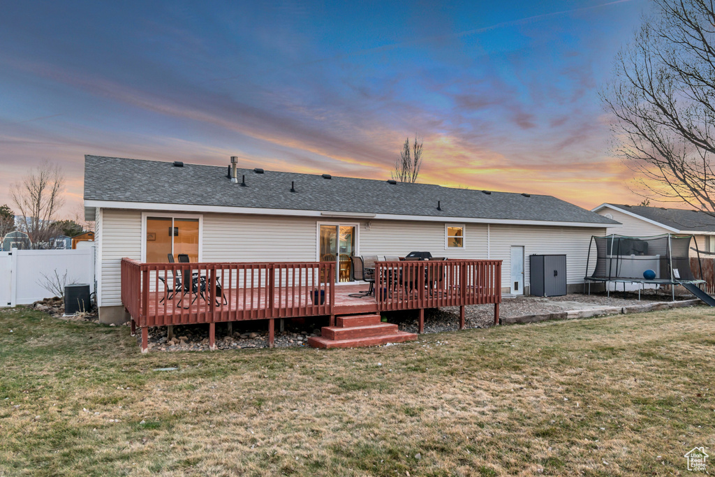 Rear view of house featuring fence, a wooden deck, a trampoline, roof with shingles, and a lawn