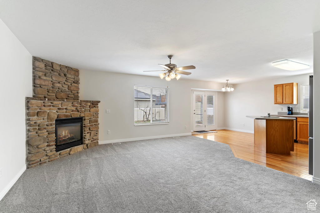 Unfurnished living room featuring a stone fireplace, light colored carpet, baseboards, and ceiling fan with notable chandelier