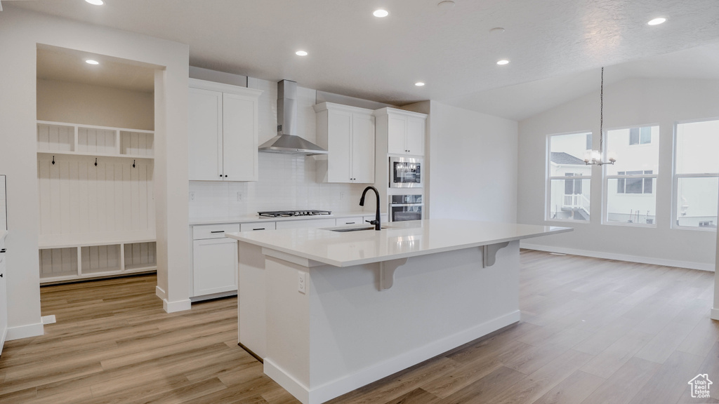 Kitchen featuring light wood finished floors, a kitchen island with sink, appliances with stainless steel finishes, a sink, and wall chimney range hood