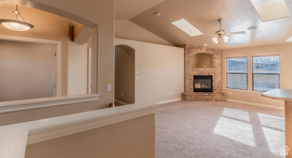 Unfurnished living room with a brick fireplace, baseboards, ceiling fan, vaulted ceiling with skylight, and light carpet