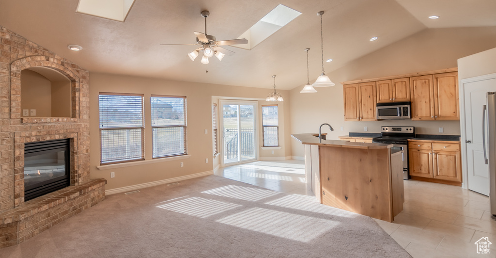 Kitchen with light brown cabinetry, a skylight, an island with sink, a brick fireplace, and appliances with stainless steel finishes