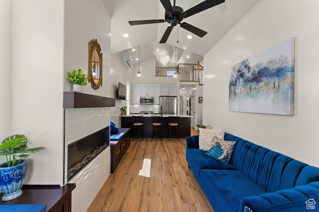 Living room featuring high vaulted ceiling, a tile fireplace, light wood finished floors, a ceiling fan, and an AC wall unit
