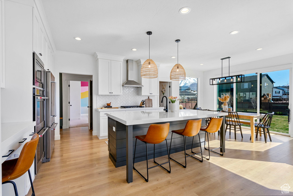 Kitchen with light countertops, white cabinets, light wood finished floors, wall chimney exhaust hood, and a kitchen breakfast bar