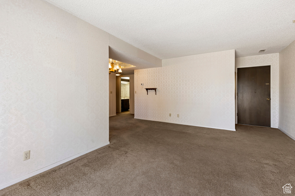 Carpeted spare room featuring a textured ceiling, wallpapered walls, baseboards, and a chandelier