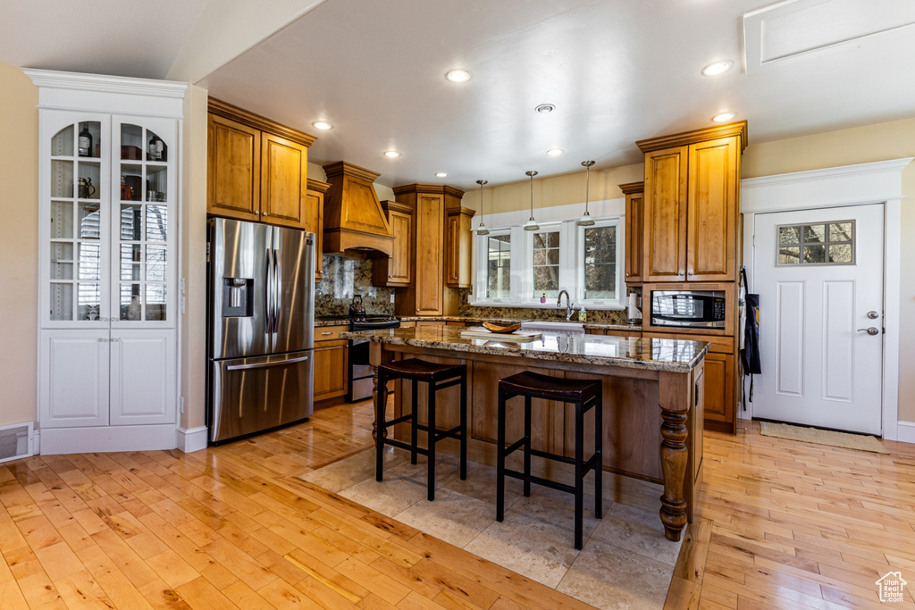 Kitchen with stainless steel appliances, custom range hood, brown cabinetry, a kitchen breakfast bar, and light wood-style flooring