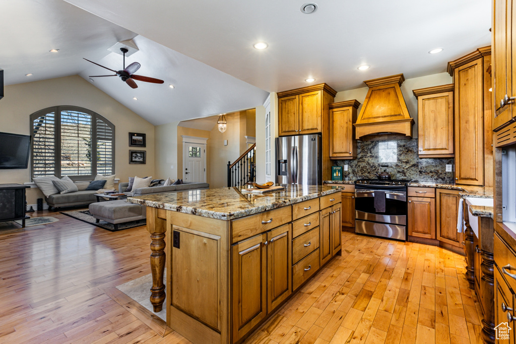 Kitchen featuring appliances with stainless steel finishes, custom range hood, a ceiling fan, brown cabinets, and light wood-style flooring