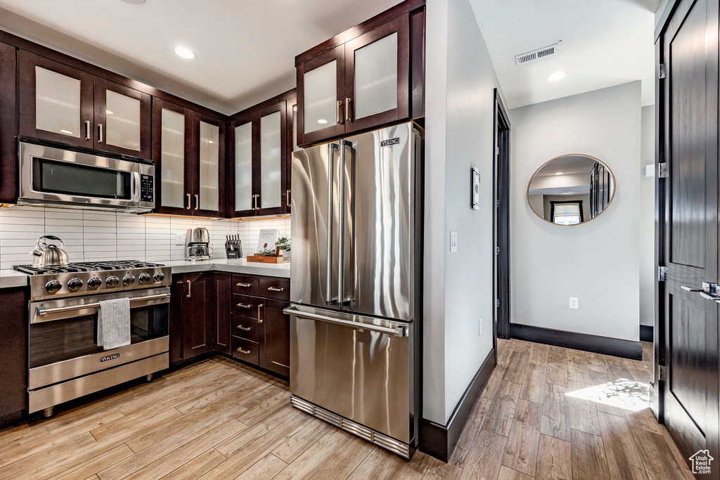 Kitchen with dark brown cabinetry, backsplash, high end appliances, visible vents, and light wood finished floors