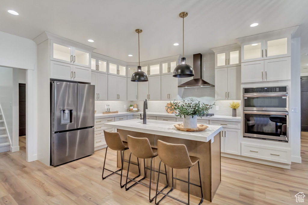 Kitchen featuring light countertops, a sink, wall chimney range hood, a kitchen island with sink, and appliances with stainless steel finishes