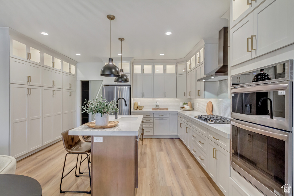 Kitchen with light countertops, wall chimney exhaust hood, light wood-type flooring, stainless steel appliances, and decorative backsplash