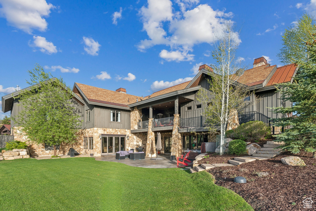 Rear view of house featuring a balcony, an outdoor living space, stone siding, a lawn, and a patio