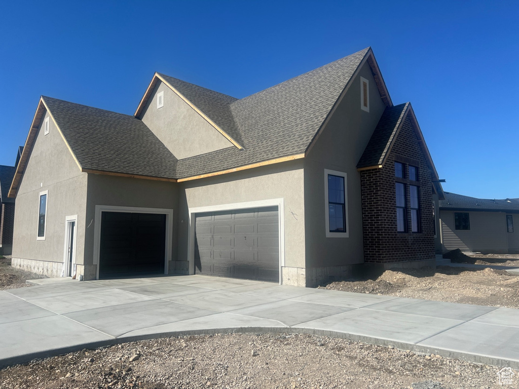View of property exterior with a shingled roof, an attached garage, brick siding, stucco siding, and concrete driveway