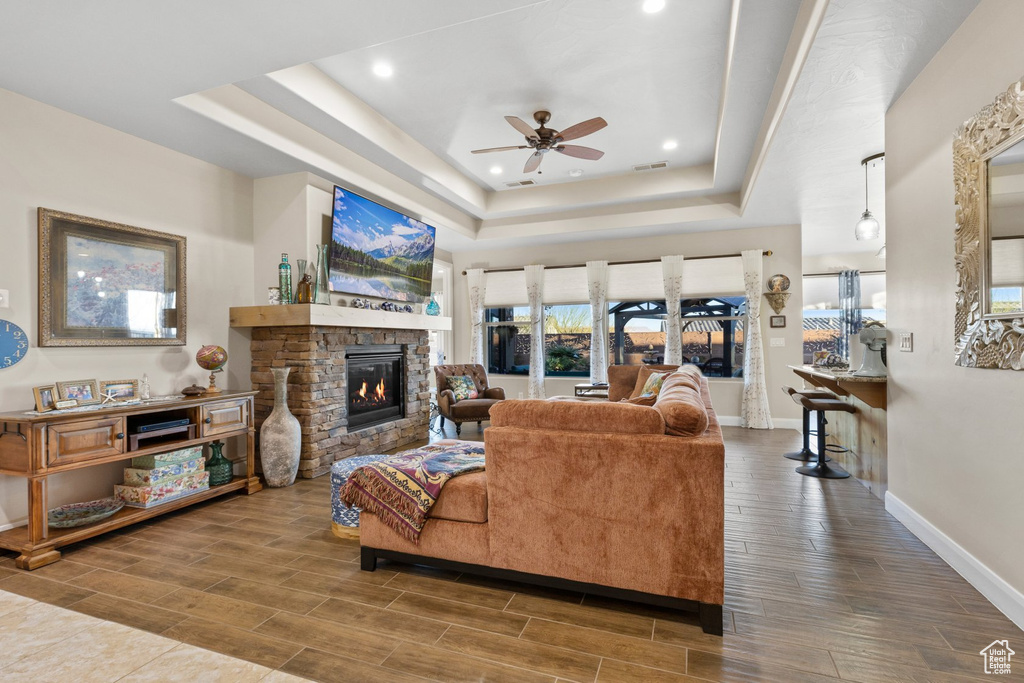 Living area featuring ceiling fan, a tray ceiling, a stone fireplace, baseboards, and wood finished floors