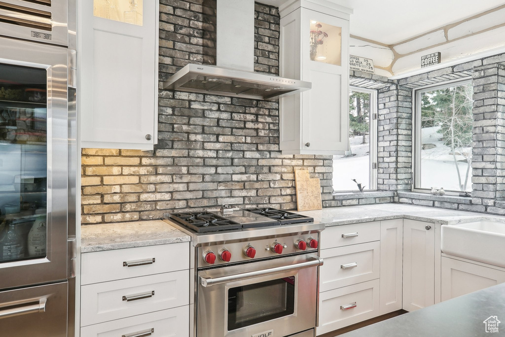 Kitchen with appliances with stainless steel finishes, wall chimney exhaust hood, white cabinets, glass insert cabinets, and light stone countertops