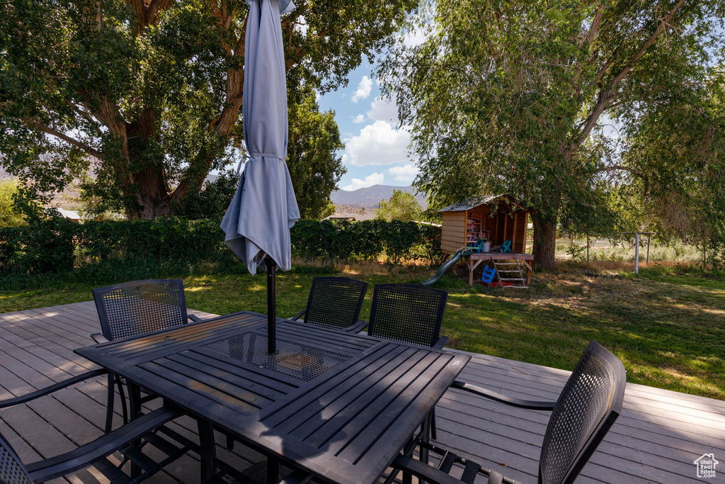 Wooden deck featuring outdoor dining area, a lawn, a playground, a mountain view, and a shed