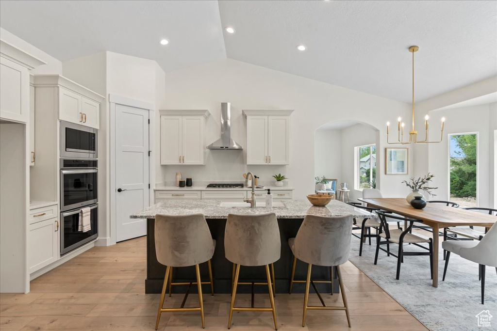 Kitchen featuring wall chimney exhaust hood, arched walkways, a center island with sink, light wood-style flooring, and a breakfast bar