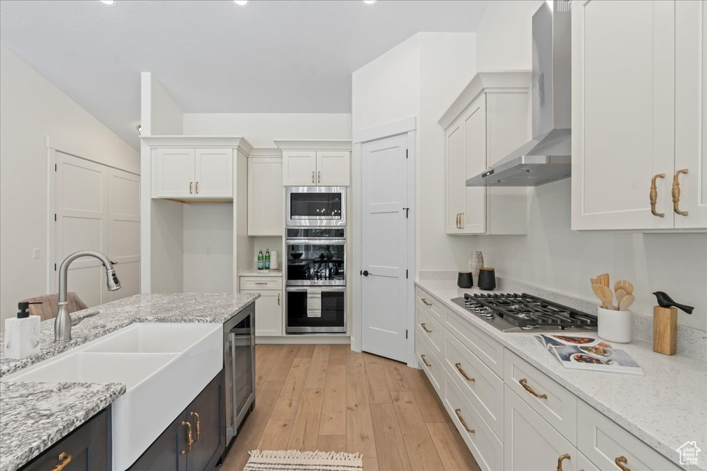 Kitchen featuring appliances with stainless steel finishes, a sink, wall chimney range hood, white cabinets, and light wood-style flooring