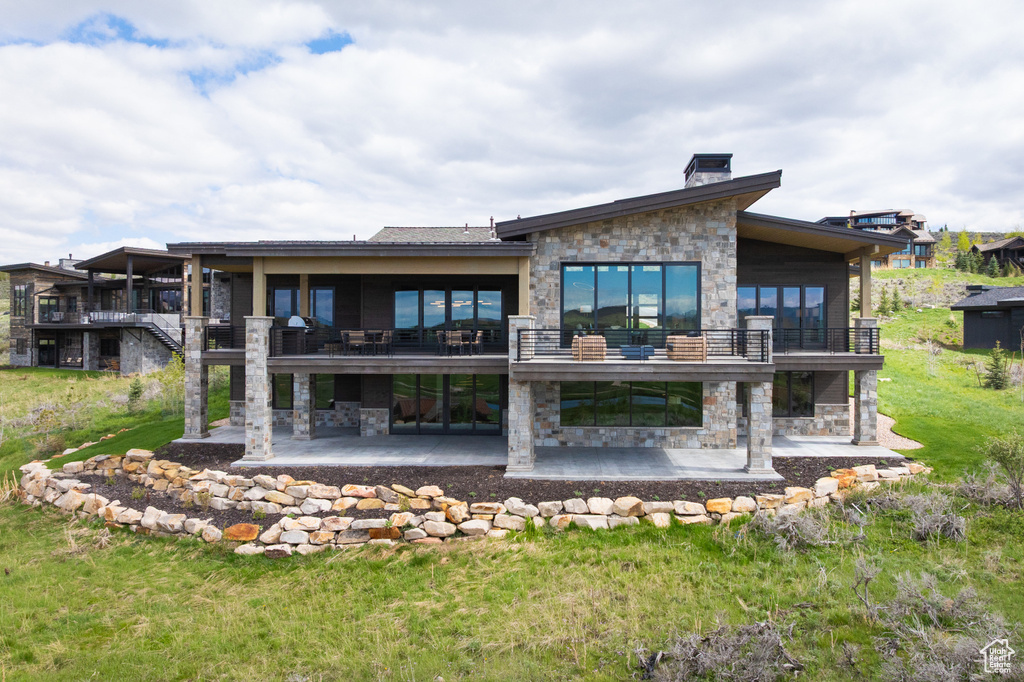 Rear view of house with stone siding, a patio, a balcony, a chimney, and a yard