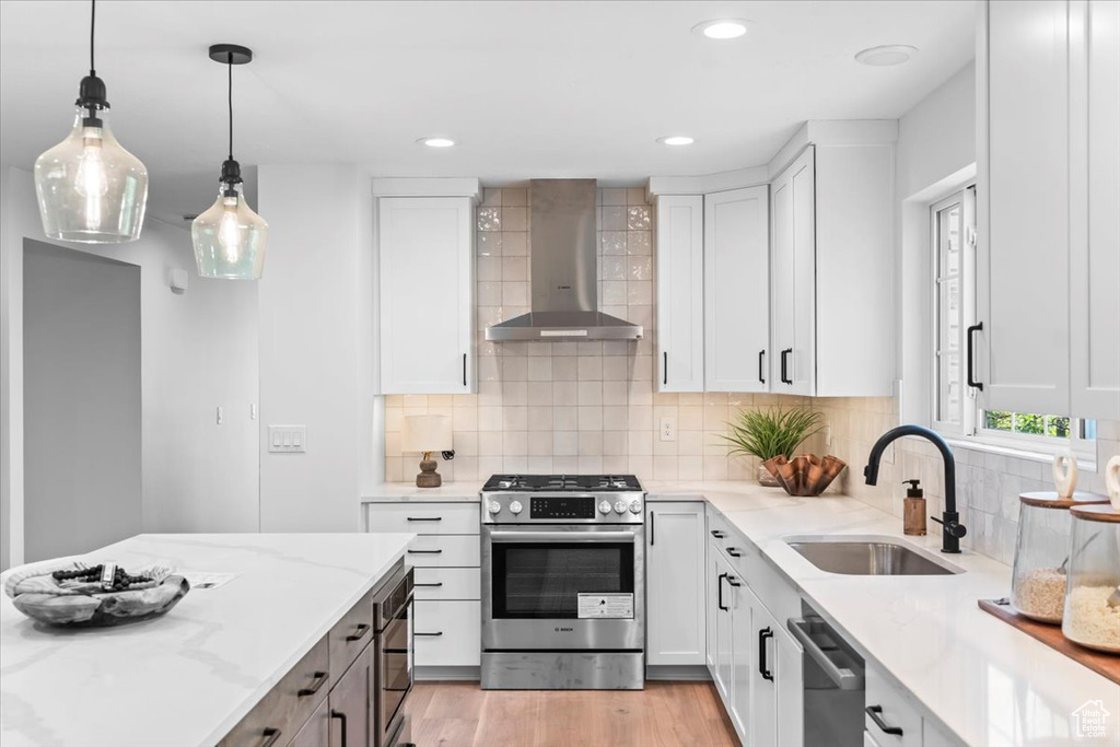 Kitchen featuring a sink, wall chimney exhaust hood, stainless steel appliances, white cabinets, and light stone countertops