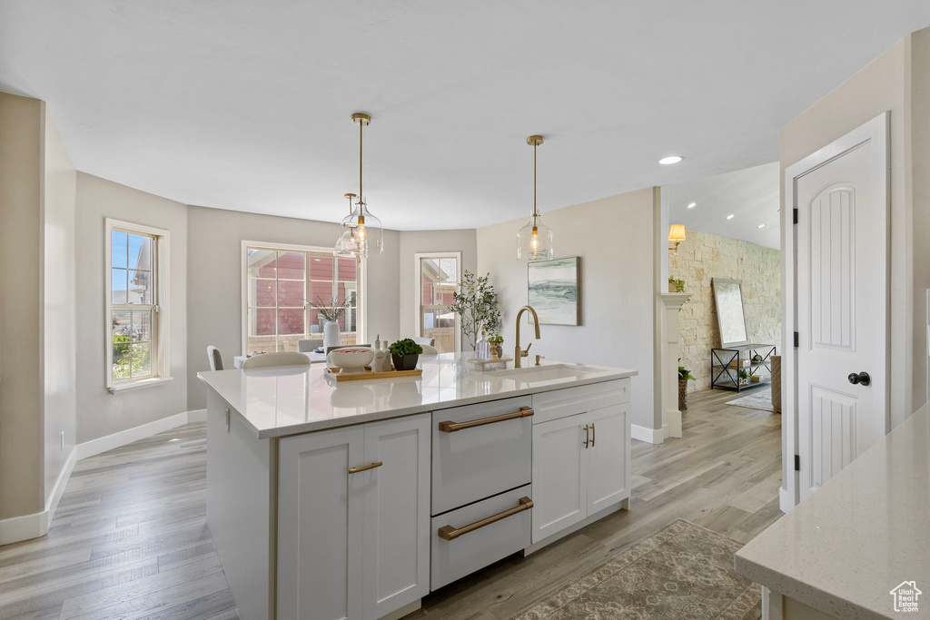 Kitchen featuring a sink, baseboards, a center island with sink, white cabinets, and light wood-style flooring